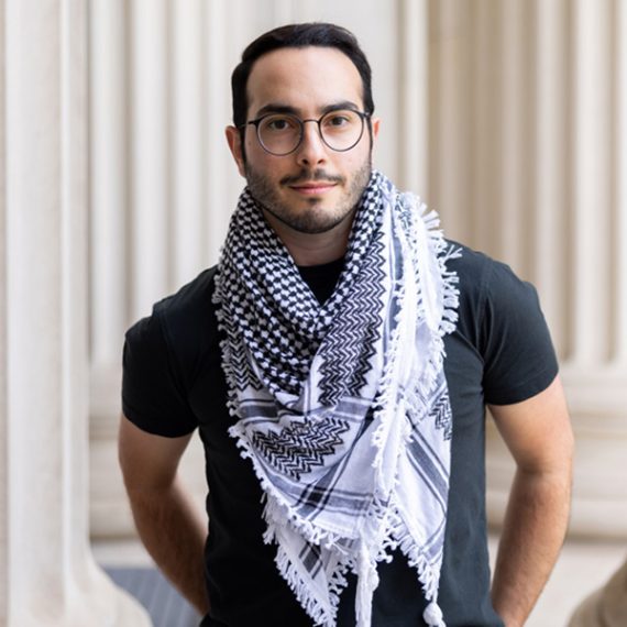 Ubadah Sabbagh stands among ivory pillars looking at camera.