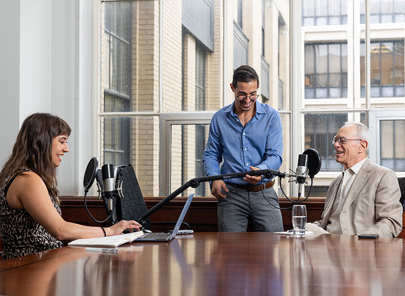 Woman interviews man at a table while another man adjusts microphone. 