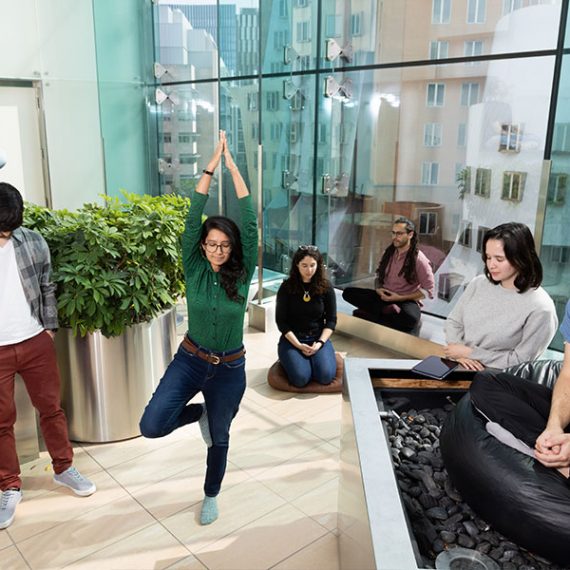 Seven people practicing different forms of mindfulness in a large room with floor to ceiling windows.