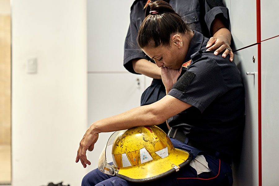 Side view of woman in uniform sitting on bench in fire station locker room with helmet in lap, head down, and eyes closed.